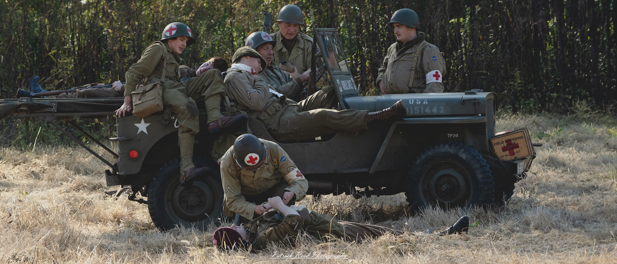 "Image of a World War II-era medic jeep ambulance, showcasing a red cross symbol prominently displayed on its side. The jeep is parked on a grassy field, with the canvas top up, providing shelter for the wounded soldiers inside. Through the open back, you can see injured personnel receiving care from a medic, highlighting the urgency and bravery of wartime medical assistance. The jeep’s weathered exterior reflects its service in the field, and the scene captures the critical role of medics in tending to the injured during the chaos of battle."